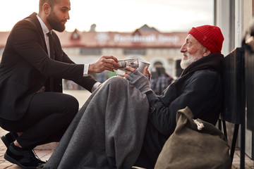Decent rich man in tuxedo hunkered down to homeless and give money donation, one dollar bill to...