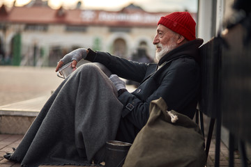 Side view on beggar male on street begging, waiting help by citizens. Senior homeless man in old...