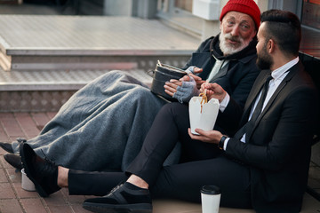 Handsome businessman in suit sitting on floor with homeless man together, listen to his story of life. Contrast people, rich and poor, but doesn't matter