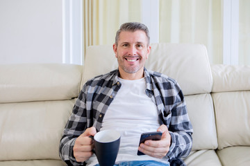 Smiling man holds a cup of tea and phone on sofa