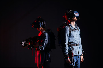 Man and woman wearing special goggles for vr futuristic games, stand back to each other. Special virtual weapons in hands. Isolated over black background