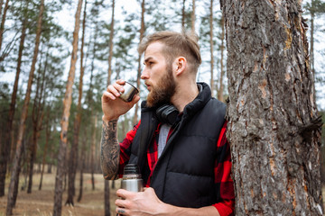 Man with beard taking rest in forest after walk, is drinking hot coffee.