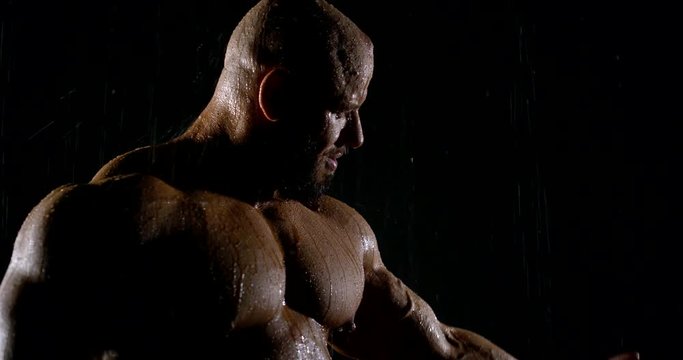 big muscular bald man in the shower. he flexes his arm and tightens his bicep. drops of water trickle down his body. side view. dark key