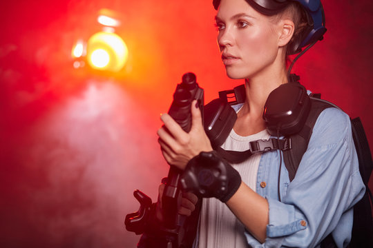 Young caucasian female playing video game under neon red light, isolated smoky background. Woman holding vr gun look side