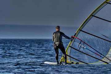 Naklejka premium A male athlete is interested in windsurfing. He moves on a Sailboard on a large lake on an autumn day.