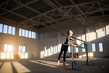 Fototapeta premium Pleasant young female athlete training at ballet bar, standing on tiptoe, closing eyes with gentle smile, expresses pleasure, shot from below, in professional sports hall