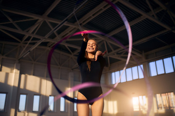 Charming young girl with closed eyes and bright smile dancing with violet ribbon in sports hall,...