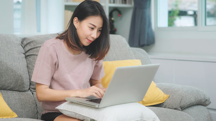 Young business freelance Asian woman working on laptop checking social media while lying on the sofa when relax in living room at home. Lifestyle latin and hispanic ethnicity women at house concept.