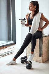 Long-haired fitness female in sportswear sit with plastic bottle after fitness. Have rest, tired. Towel on shoulders.