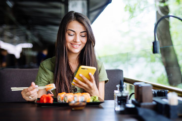 Beautiful girl eats sushi and looks in smartphone in cafe