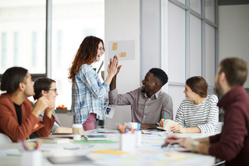 Portrait of creative business team giving high five while planning project during meeting in office, focus on smiling young woman standing by table