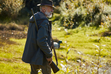 Mature man hunter in hat stand near lake with shotgun, going to hunt on wild ducks. Autumn hunting