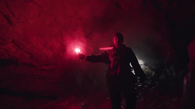 Man lighting his way inside dark cave with a burning signal flare and leaving the entrance behind him. Stock footage. Dangerous tourism inside dark grotto.