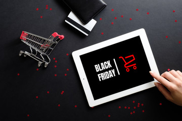 Black Friday sale concept. Woman hand using tablet with sign "Black Friday" on screen, shopping cart and debit card over black background. Flat lay, top view, overhead. © photoguns
