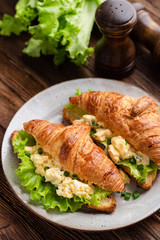 Croissant egg sandwich with green lettuce salad on plate on wooden table. Top view. Healthy tasty breakfast sandwich
