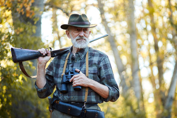 Senior hunter on birds holding gun on shoulder, straighten hat, looking away. Forest background