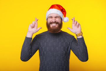 Photo of young bearded man wearing santa claus hat and standing with crossed fingers and dreaming