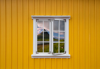 Yellow wooden house with a white window with a reflection of the Icelandic landscape