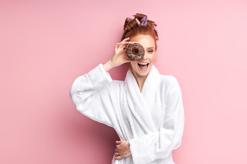 Gorgeous redhaired woman holding sweet donut, happy after shower in bathrobe and curlers on hair. Isolated over pink background - obrazy, fototapety, plakaty