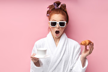 Caucasian girl after shower enjoying cup of tea with croissant. Opened mouth. Isolated over pink...