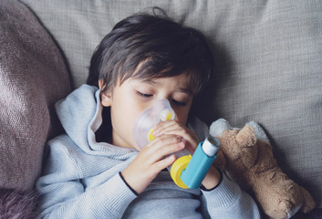 Poor boy tired from chest coughing holding inhaler mask, Child closing his eyes while using the volumtic for breathing treatment,Tried Kid having asthma allergy using the asthma inhaler - obrazy, fototapety, plakaty