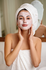 Woman with towel is applying mask on her face at home, close-up. Skin care.