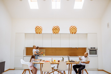 Daytime with family at home. Father busy working from home, using laptop. Mother cooking while holding baby.