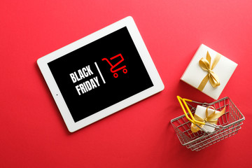 Black Friday concept. Tablet with sign "Black friday" on screen, shopping cart, gift box over red background