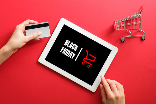 Black friday online shopping concept. Hand with debit card, tablet with "Black friday" sign on screen, shopping cart over red background.