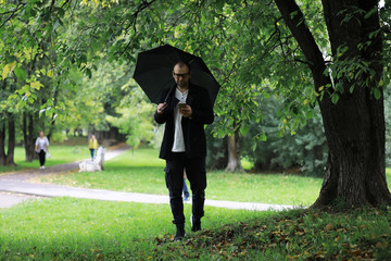 A young man in glasses walks in the park with an umbrella during the rain.