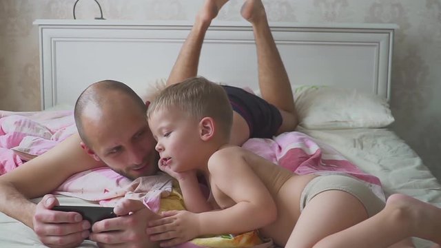 Dad and son lying on bed and using mobile phone together