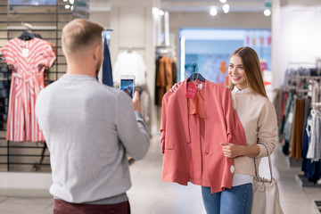 Young man with smartphone taking photo of his girlfriend holding pink jacket