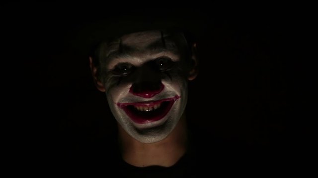 Scary clown looks at the camera and laughs terribly. A scary man in a clown makeup looks at the camera and laughs. Scary clown grimaces looking into camera .Halloween.