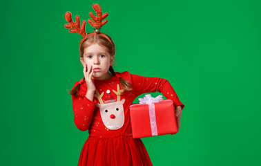 sad, surprised funny child girl in red Christmas reindeer costume with gift on green   background.