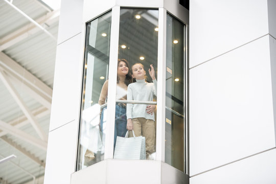 Young mother and daughter discussing something while moving upwards in elevator