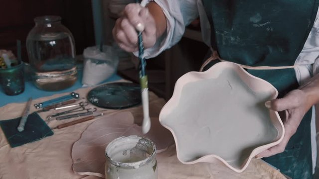professional potter decorating and painting a dish after she has baked it in the kiln. Potter applying first level of glaze on her work of art. Potter adding interesting details to a plate. Potter