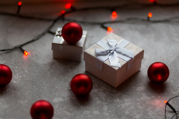 Christmas decorations on a gray background. A white with a bow is surrounded by unfocused burgundy Christmas balls, Christmas toys and red garland.