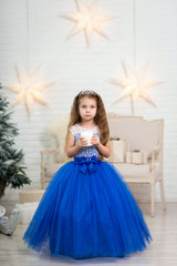 Cute little girl in a magnificent blue dress holding an artificial candle in her hands for home decoration on Christmas holidays. Child-friendly scenery