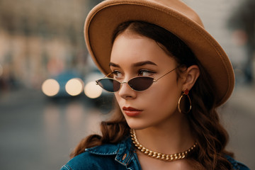 Outdoor close up fashion portrait of young elegant lady wearing beige hat, trendy sunglasses, earrings, chain necklace, denim shirt, posing in street of European city. Copy, empty space for text - obrazy, fototapety, plakaty