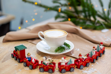 Delicious fresh festive morning cappuccino coffee in a ceramic white cup on the wooden table with decorative christmas train, red ornamentals, fireflies and spruce branches