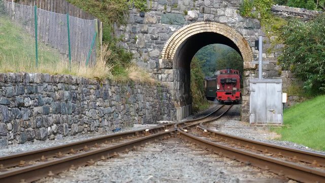 Steam engine train approaching short arch tunnel under road bridge.