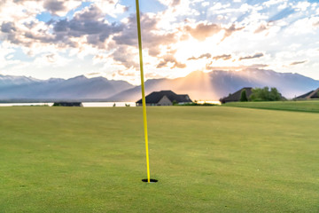 Obraz premium Yellow flagstick and hole at a golf course against lake mountain and cloudy sky