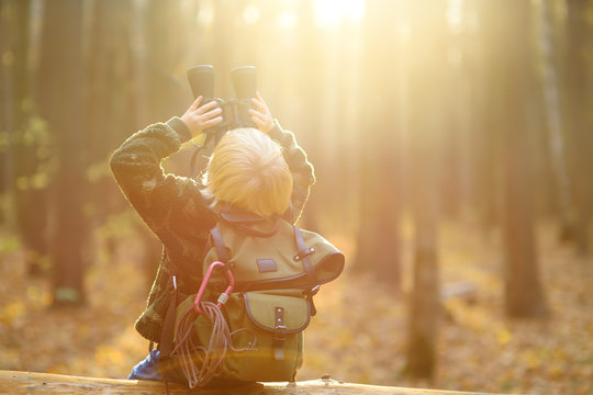 Little boy scout with binoculars during hiking in autumn forest. Child is sitting on large fallen tree and looking through a binoculars.