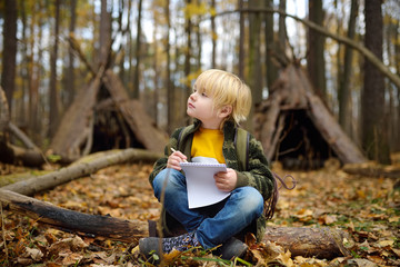 Little boy scout is orienteering in forest. Child is sitting on fallen tree and writing in the...