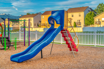 Blue slide with red stairs at a playground against homes mountain and blue sky - obrazy, fototapety, plakaty