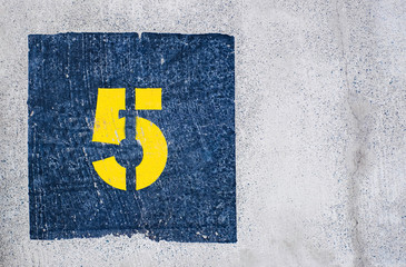 Yellow number 5, five, in dark blue square, offset on flat concrete surface.