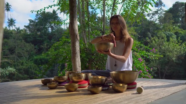 Woman playing on Tibetan singing bowl while sitting on yoga mat against a waterfall