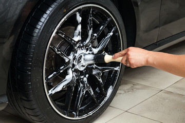 A male worker washes a black car with a special brush for cast wheels and scrubs the surface to...