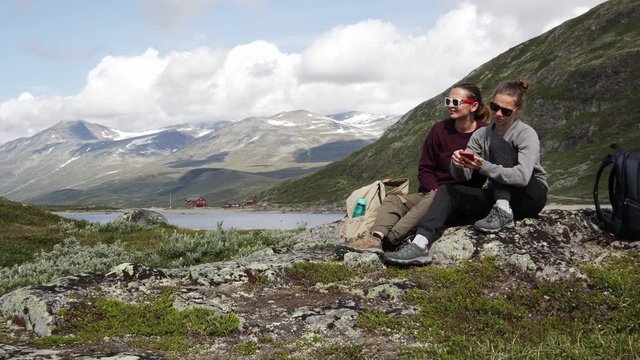 Two young women travelers relaxing with a view to a Norwegian fjord and using a smartphone. Backpacker trip to Norway. 