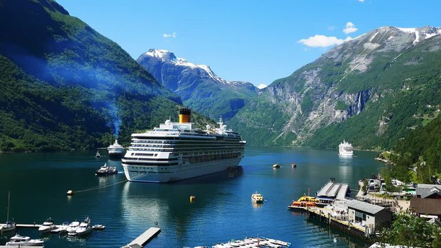 Spectacular clip of Geiranger Fjord and mountains with snow and waterfalls and cruise liners and ferry boats carrying tourists from ship to shore on a beautiful day with blue sky and fluffy clouds.
